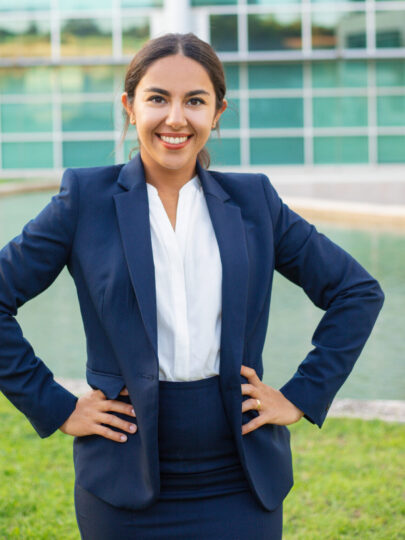 Happy confident young professional posing outside. Beautiful Latin woman wearing formal jacket, standing near office building, looking at camera and smiling. Business professional concept