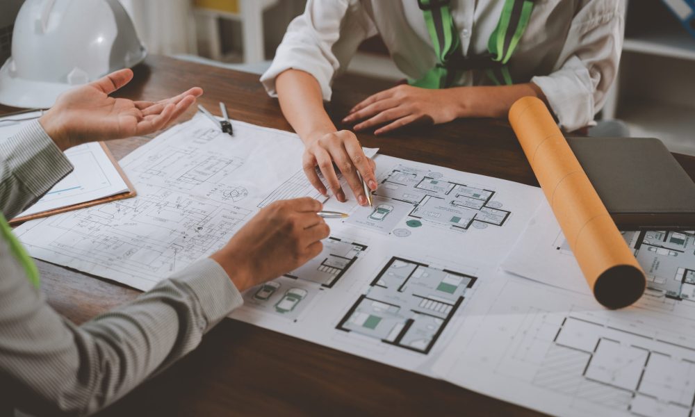 Two architects are discussing house plans in a meeting, pointing at blueprints and diagrams on a wooden table